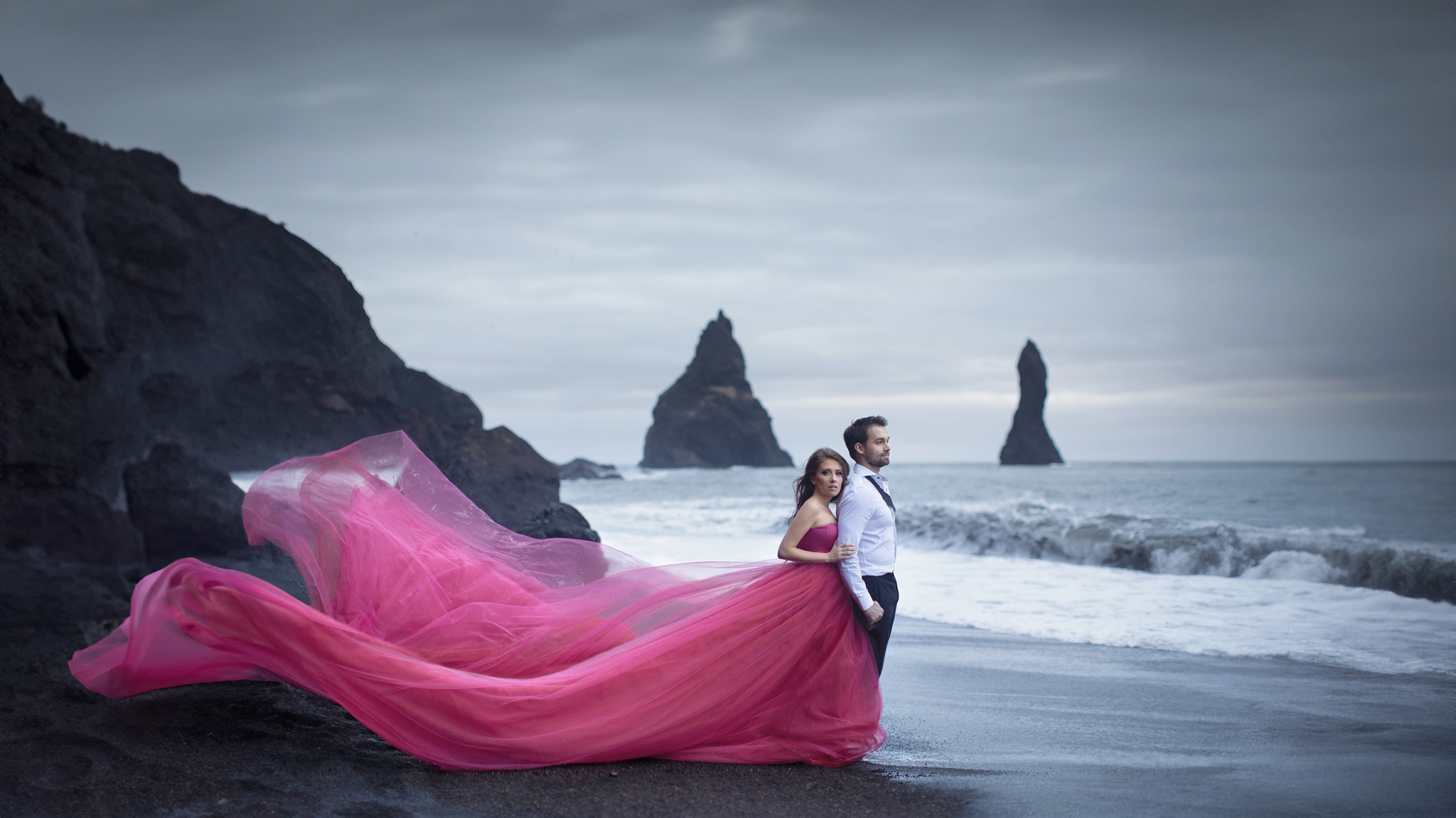 Couple standing on an Icelandic black-sand beach with sea stacks behind them
