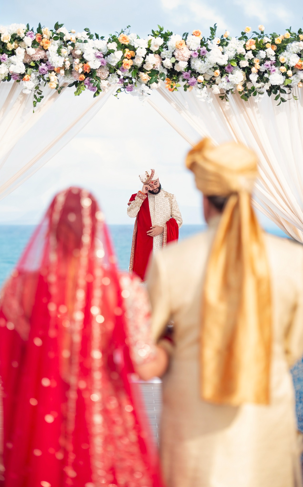 Groom reacting as the bride approaches beneath a floral ceremony canopy by the ocean